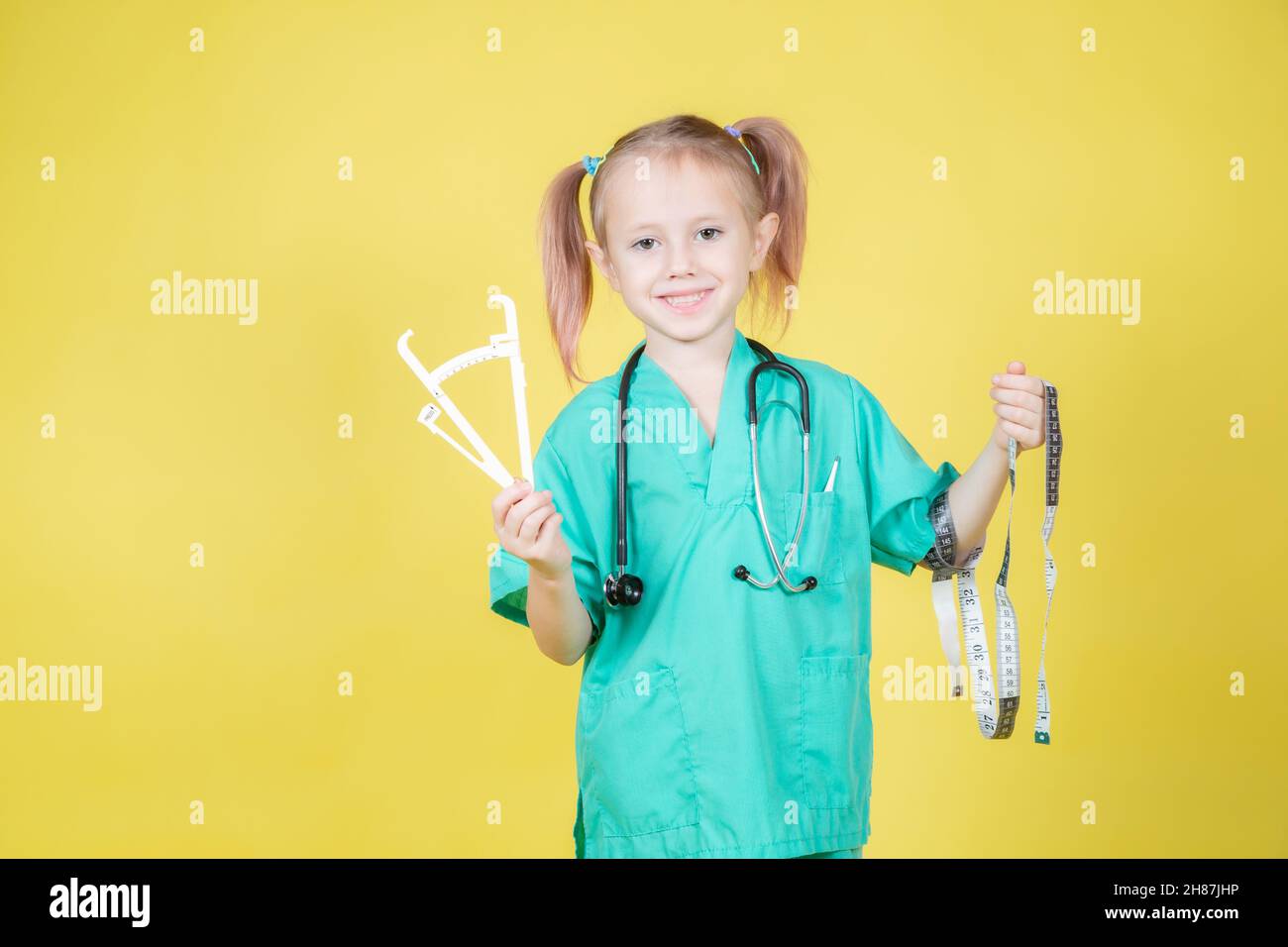 Portrait of little caucasian kid dressed in doctors green coat holds caliper and tape measure in hand isolated on yellow Stock Photo