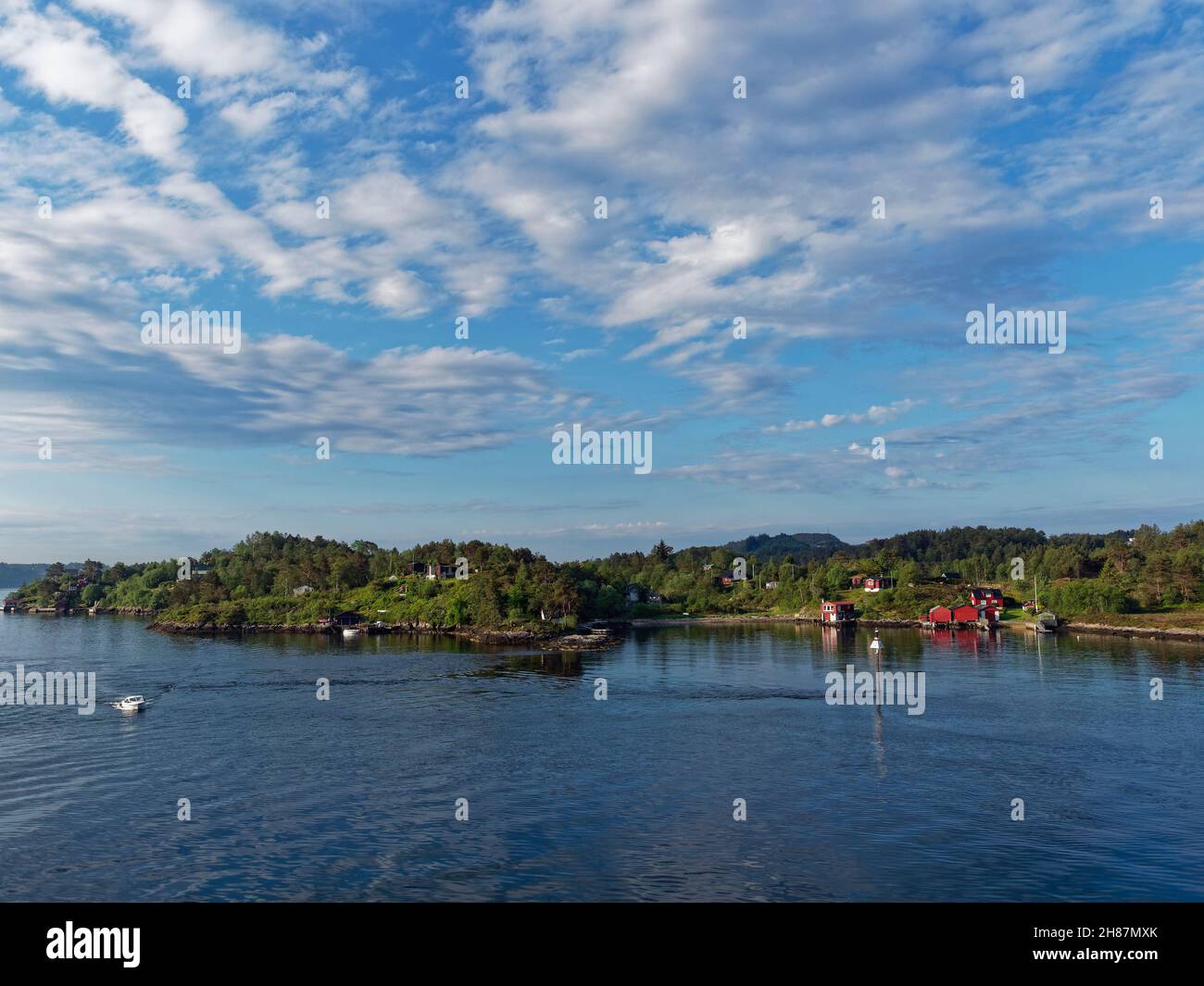 A Small Motorboat cruising past the forested coastline with scattered Houses and Cabins in Bergen Fjord on a bright Summers Morning in June. Stock Photo