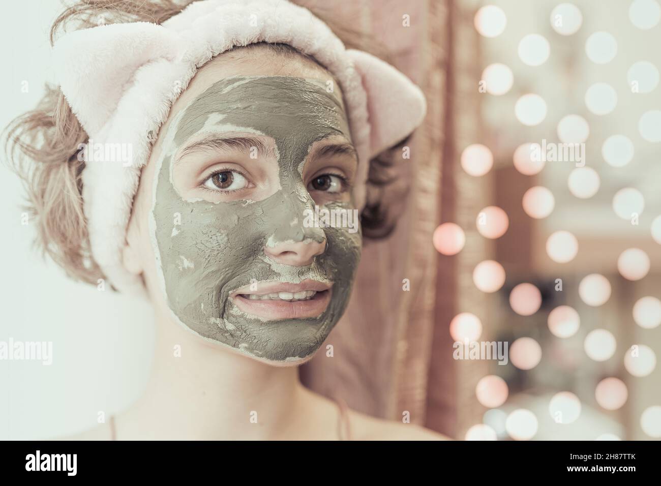 Caucasian model girl teenager in towel on her head wearing cosmetic mask made of green healing clay on blurred background of lights of festive garland Stock Photo