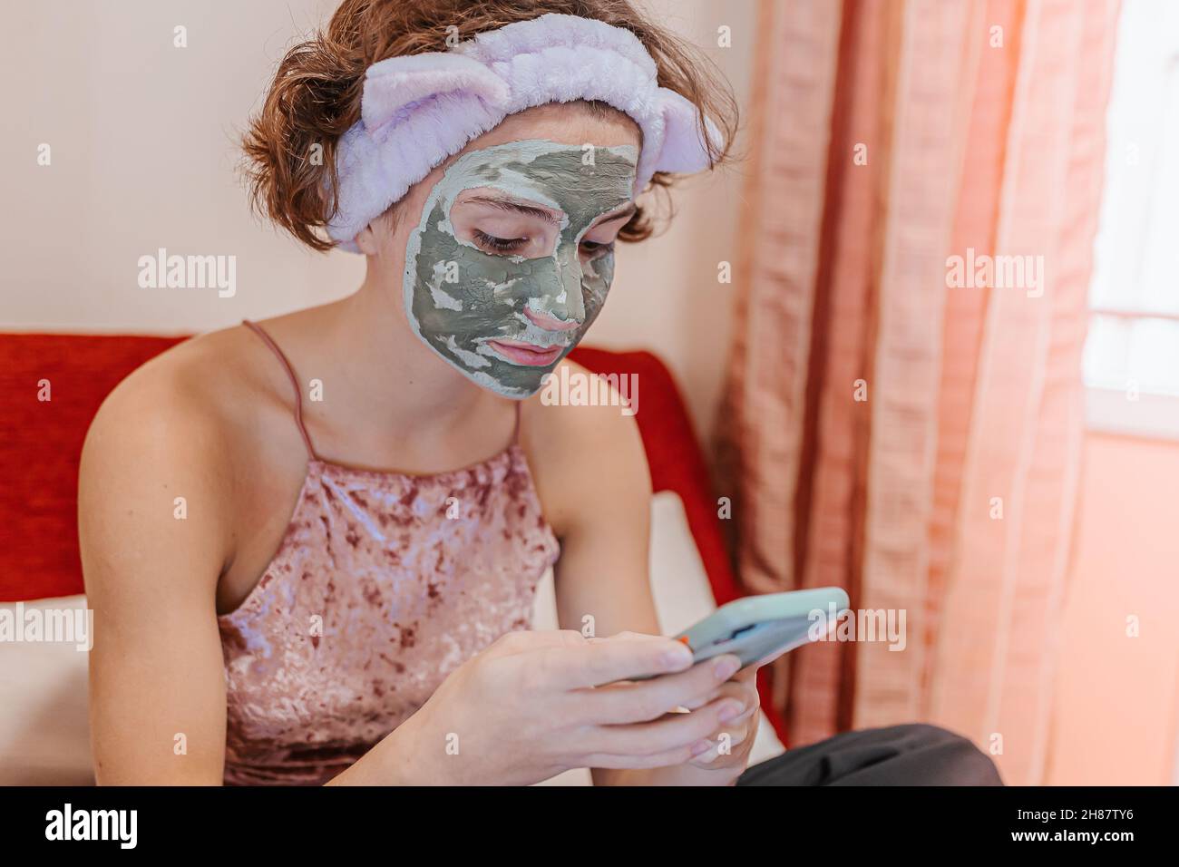 teenage girl brown curly hair clay cosmetic mask on her face takes care of her face at home and communicates online writes message her girlfriend whil Stock Photo