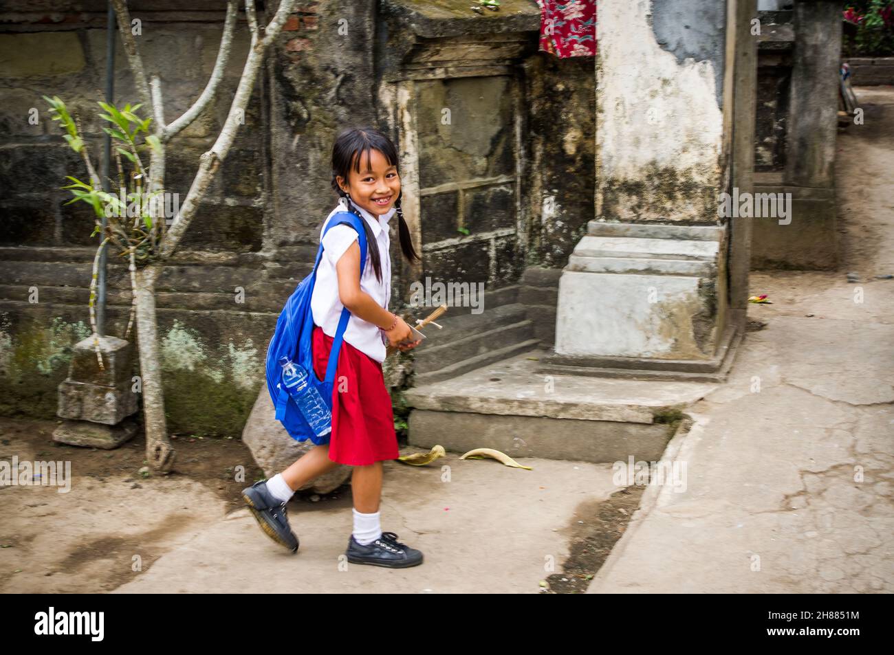 An Indonesian student is coming home from school. She is smiling and wearing a red skirt and a white shirt. Stock Photo