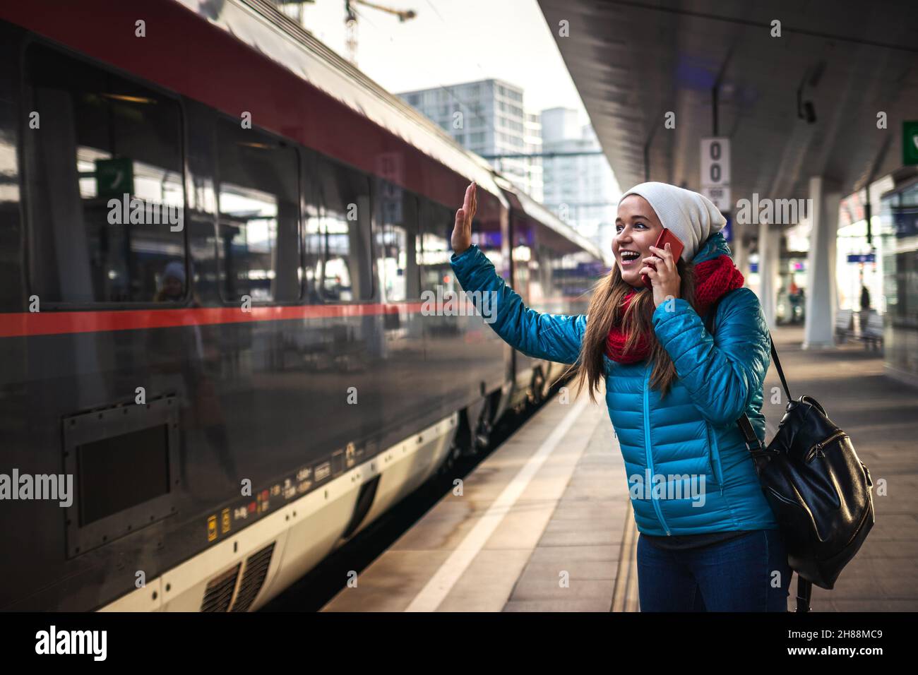 I can see you! Happy woman waving at passenger in the arriving train and talking to mobile phone. Smiling young woman looking to train at station Stock Photo