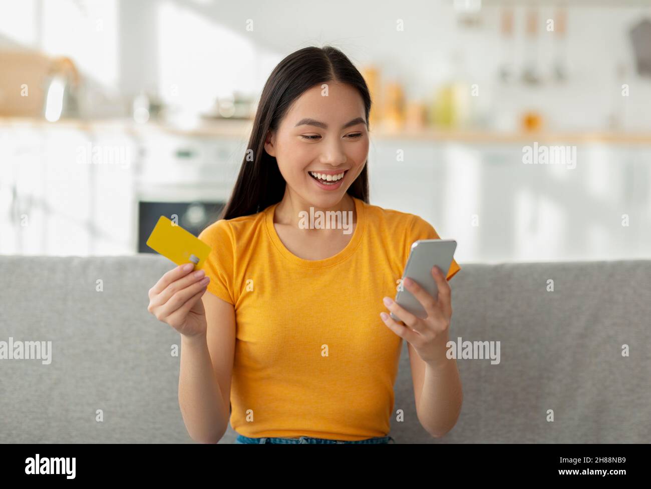 Online payment concept. Excited asian lady holding credit card and smartphone, sitting on sofa, making internet purchase Stock Photo