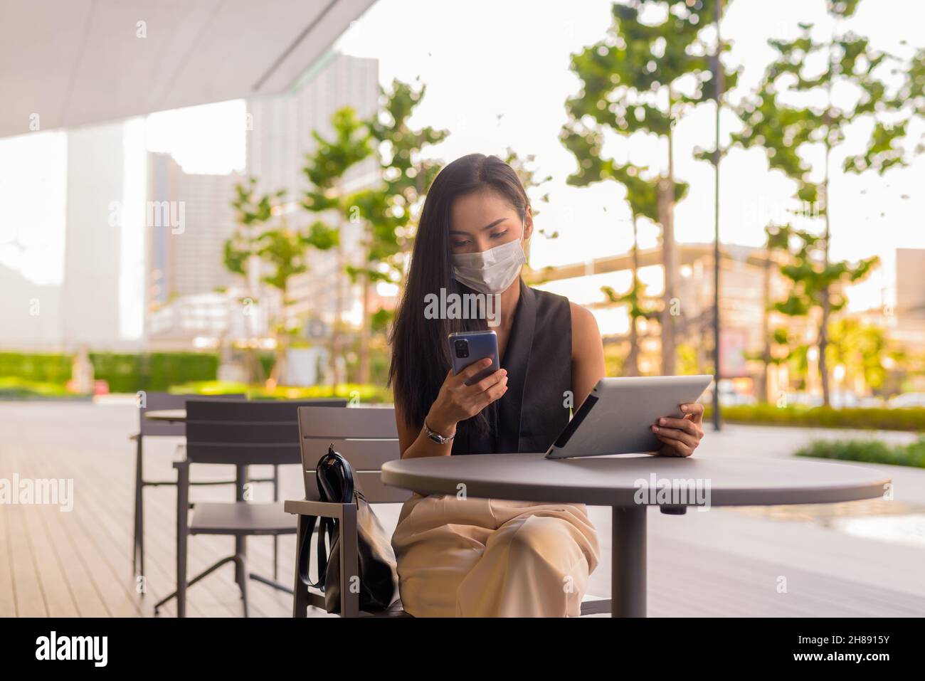 Woman sitting outdoors at coffee shop restaurant social distancing and wearing face mask to protect from covid 19 while using phone and digital tablet Stock Photo