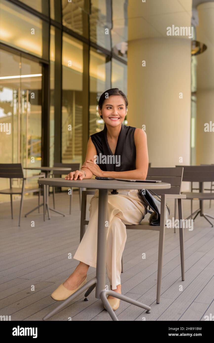 Full length portrait of beautiful Asian woman sitting outdoors at coffee shop restaurant while smiling and thinking Stock Photo