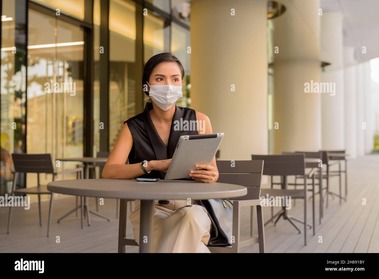 Woman sitting outdoors at coffee shop restaurant social distancing and wearing face mask to protect from covid 19 while using phone and digital tablet Stock Photo