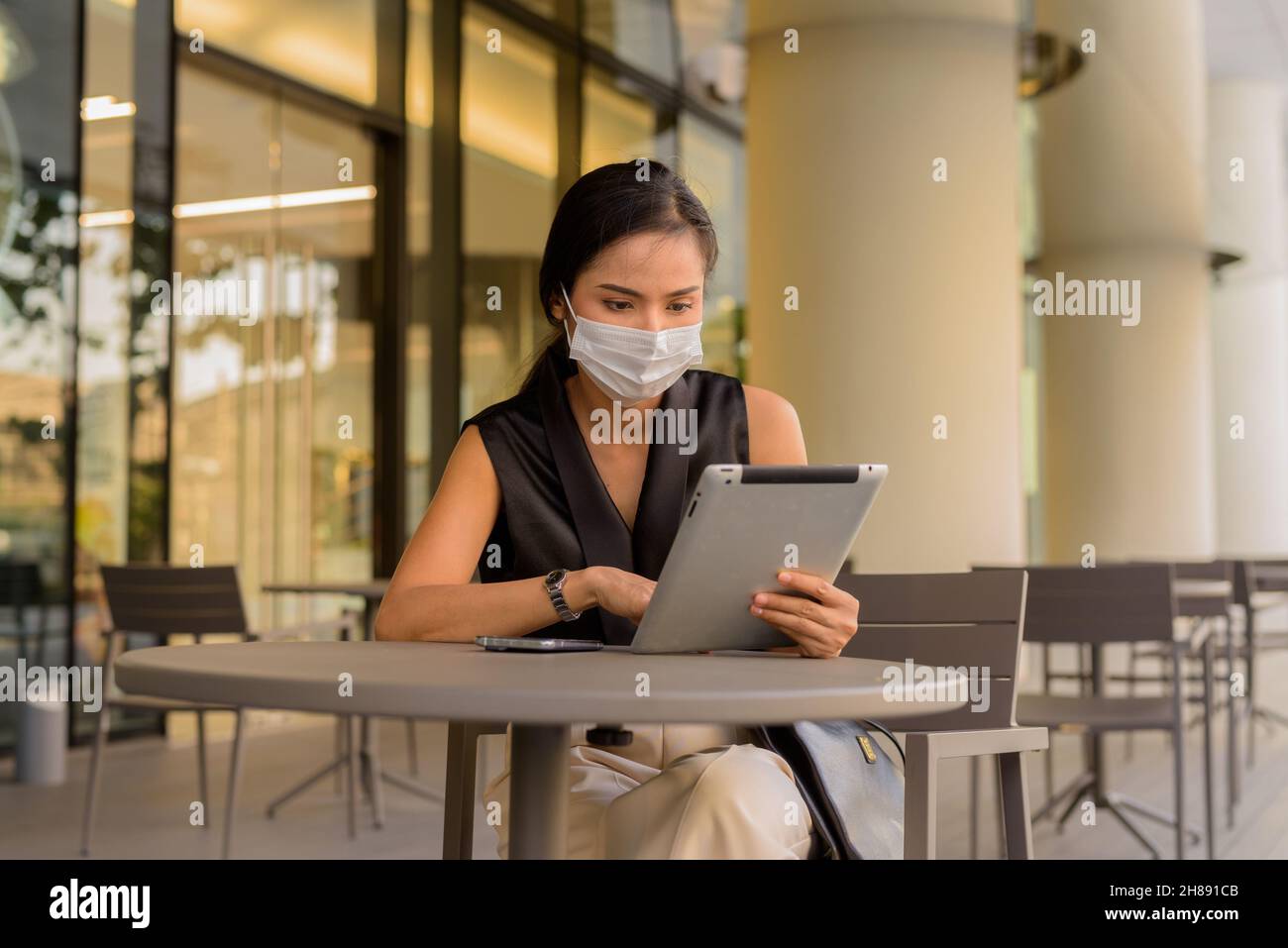 Woman sitting outdoors at coffee shop restaurant social distancing and wearing face mask to protect from covid 19 while using phone and digital tablet Stock Photo