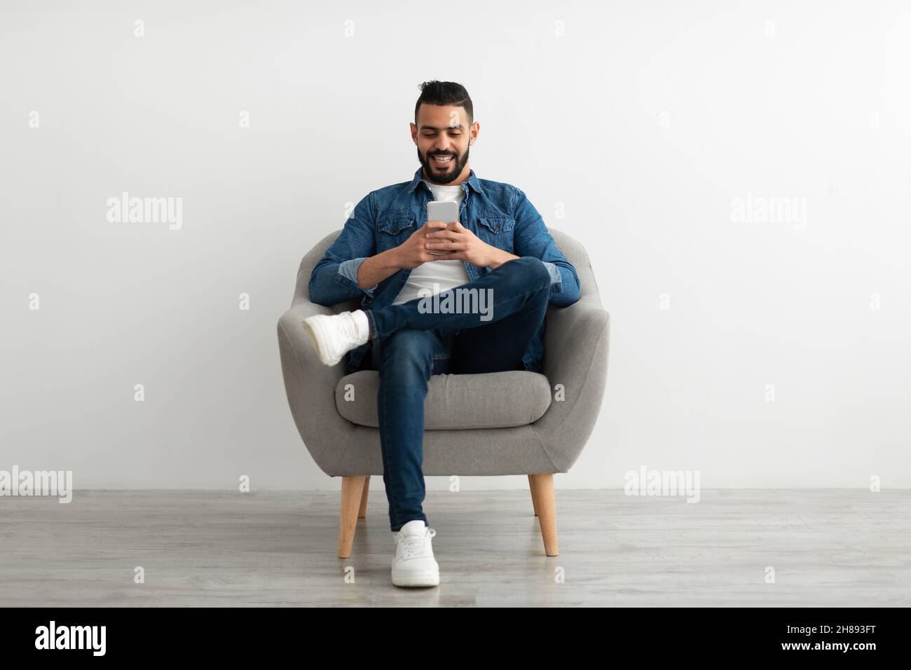 Cheerful young Arab man using smartphone, having online chat, sitting in armchair, browsing social media Stock Photo