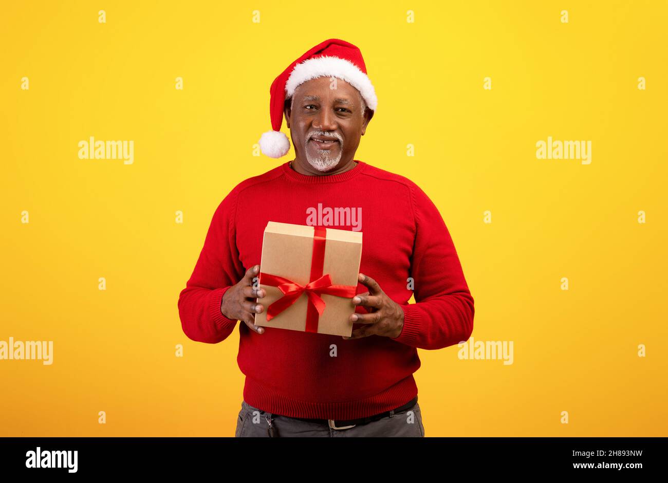 Positive senior black man holding wrapped Xmas gift box, wearing Santa hat and red sweater on orange background Stock Photo