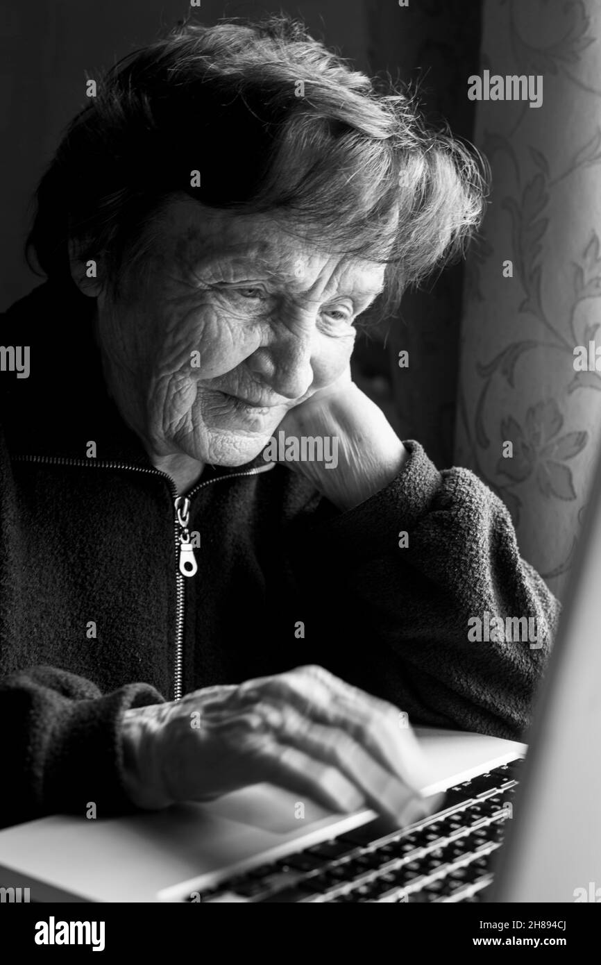 An old woman is typing on a laptop in her home. Black and white photo. Stock Photo
