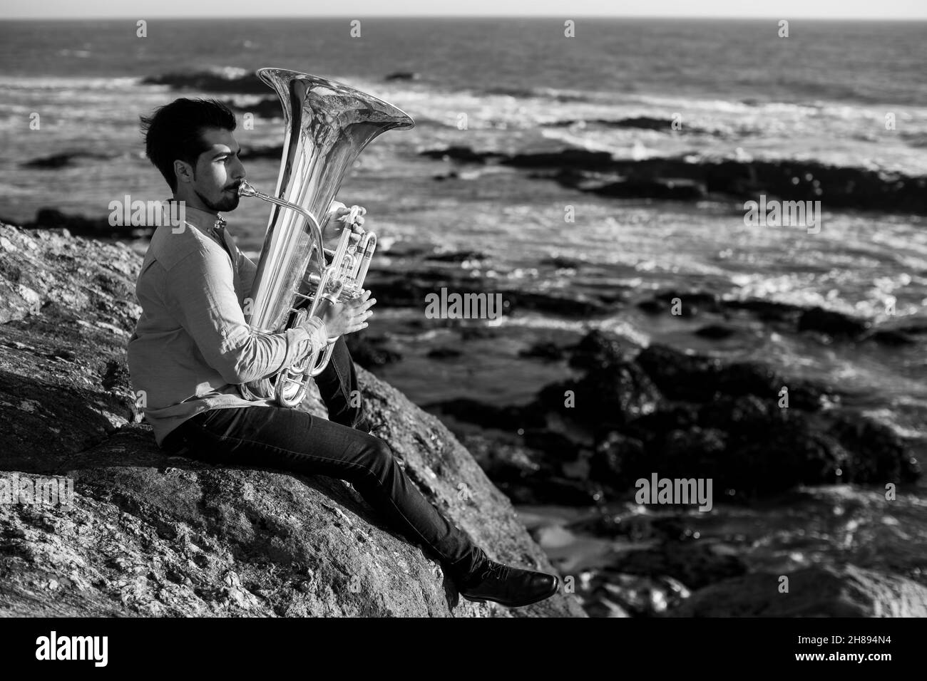 Musician sitting on the rocks with a tuba on the ocean shore. Black and white photo. Stock Photo