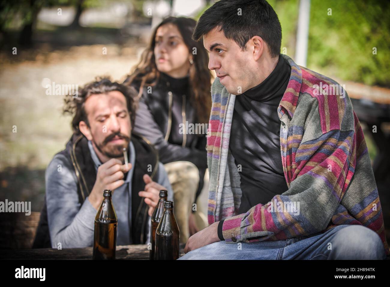 Group of female and male young friends drinks beer from bottle and smoking cannabis marijuana ganja or hashish joint in public park outdoor. Young peo Stock Photo