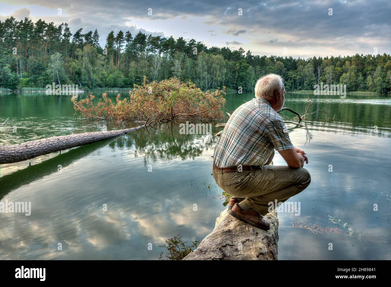 In the last light of the setting sun an old man sits on a log on a lonely lake and enjoys the silence and quiet of the evening hour in nature. Stock Photo