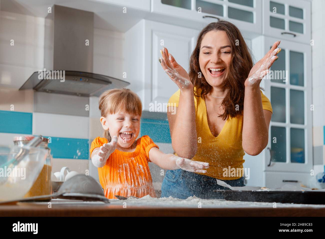 Happy laughing mother and daughter preparing baking throw flour in kitchen together. Family mom and child kid enjoys common leisure time cooking food Stock Photo