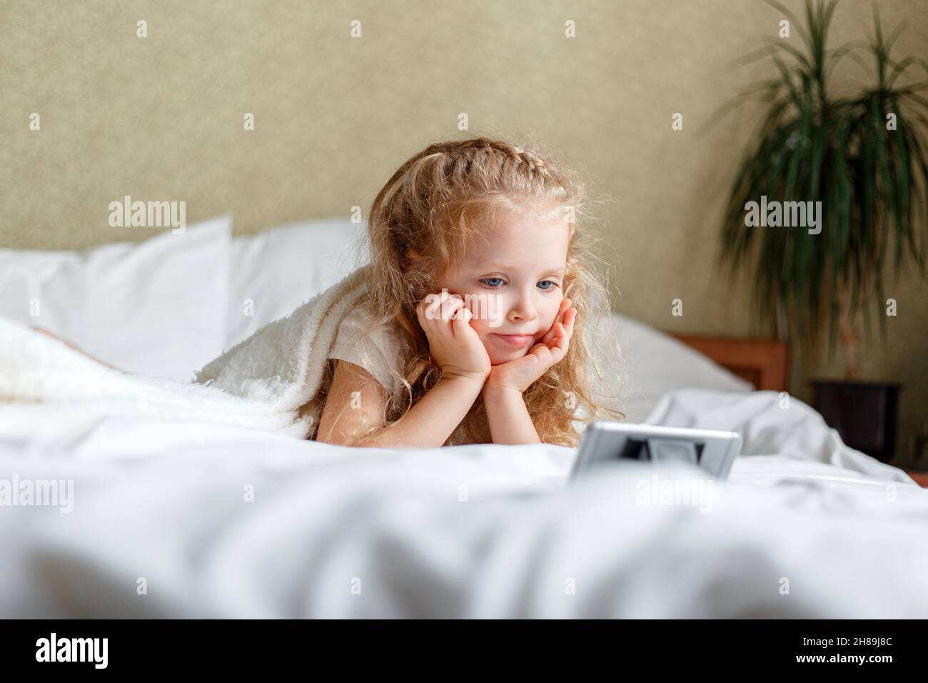 Little beautiful caucasian girl preschooler lies in bed watching video or cartoons using smartphone. Kid child girl spend leisure time watching videos Stock Photo