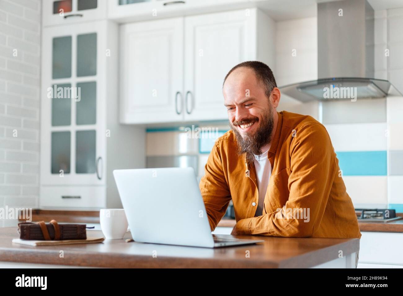 Young man working using laptop in kitchen in home interior. Smiling Freelancer works remotely from home. Happy man owner is doing paperwork using Stock Photo
