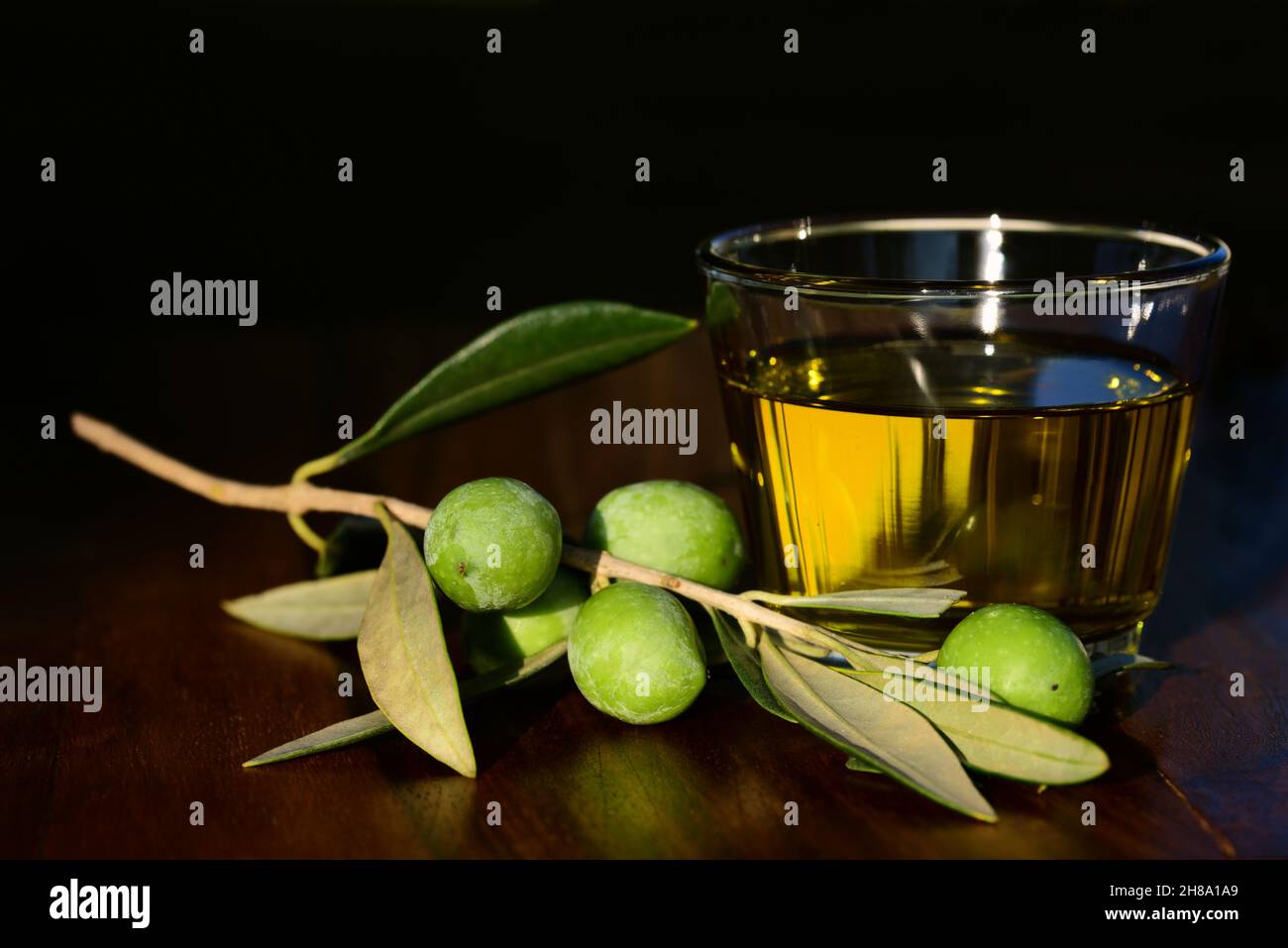 a glass with olive oil stands on a wooden table against a dark background. Next to it is a branch with olives and leaves on it Stock Photo