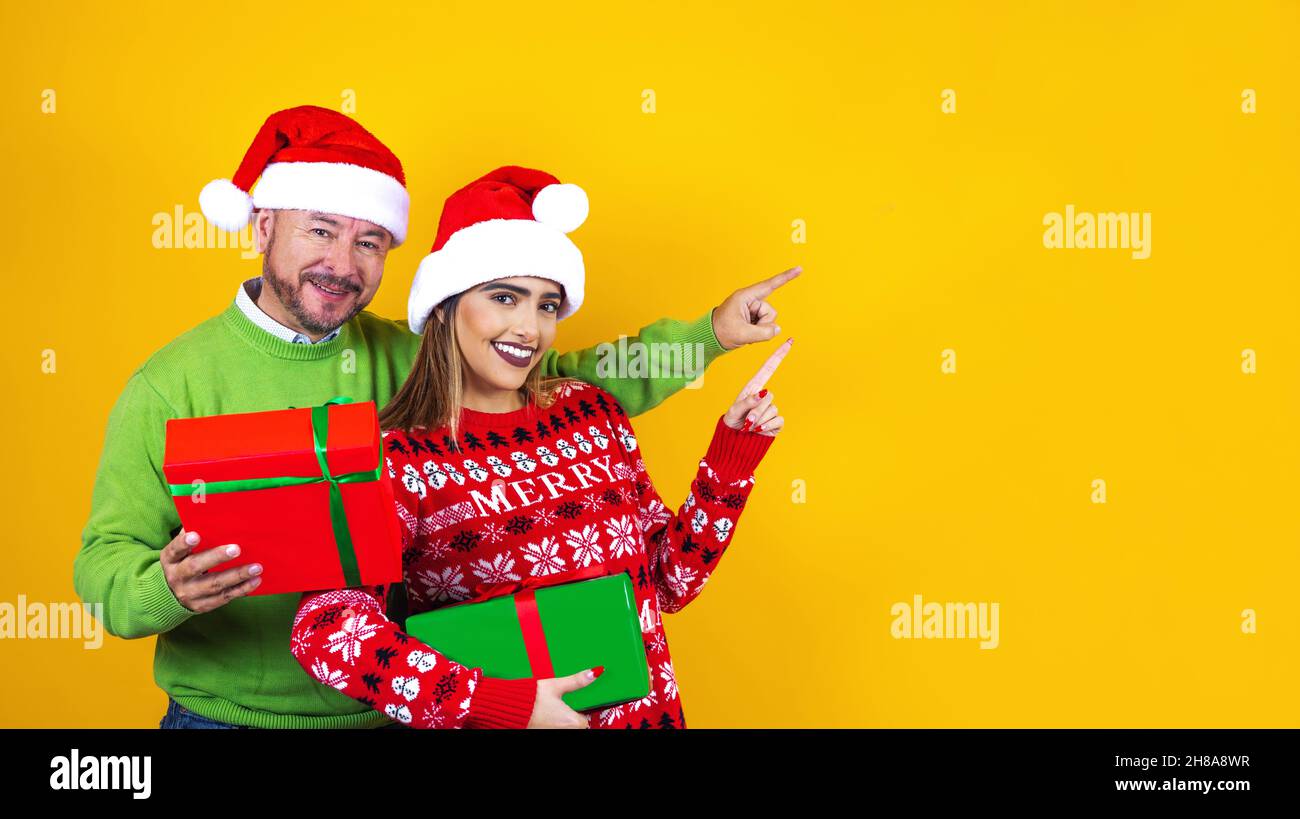 latin father and daughter in red sweater Christmas santa hat holding christmas presents pointing copy space on yellow background in Mexico Latin Ameri Stock Photo