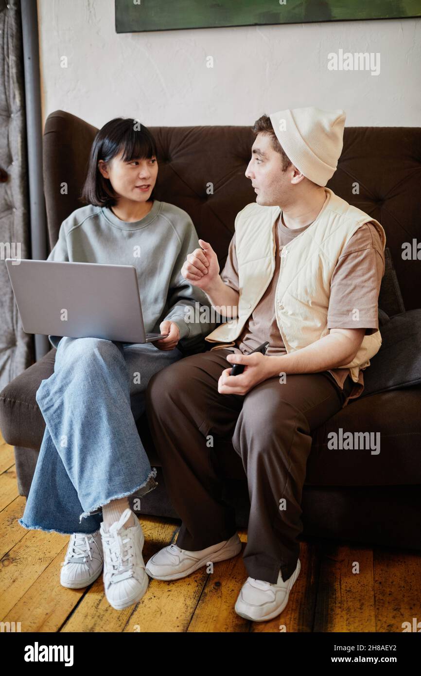 Two students sitting on sofa using laptop and discussing online education together in the room Stock Photo