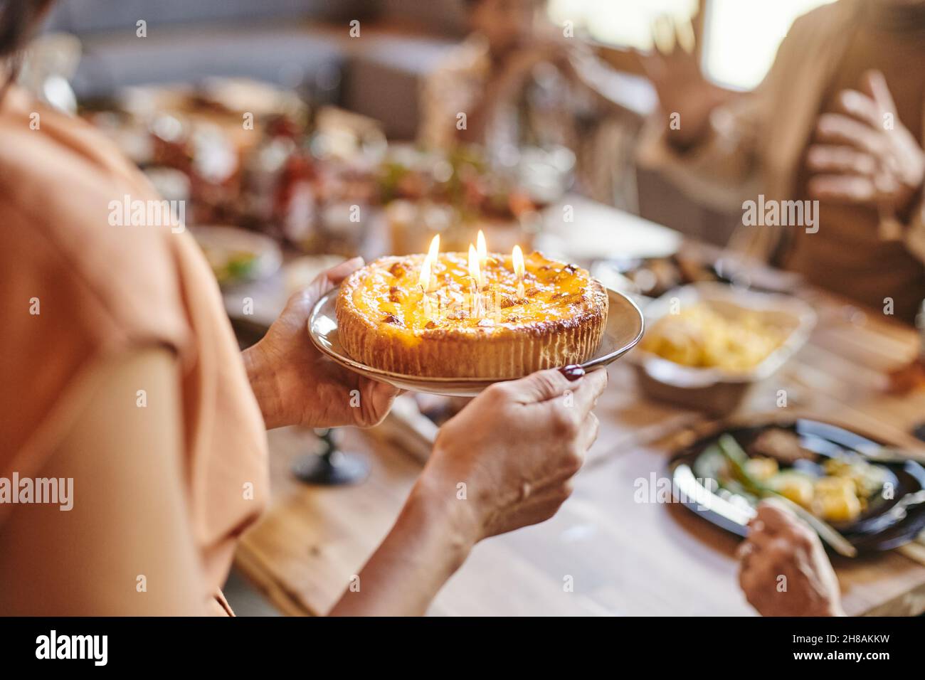 Hands of young woman holding plate with homemade birthday cake with several burning candles while standing by festive table Stock Photo