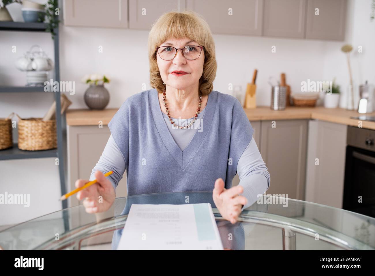 Blond mature teacher in casualwear and eyeglasses explaining something to students during online lesson Stock Photo