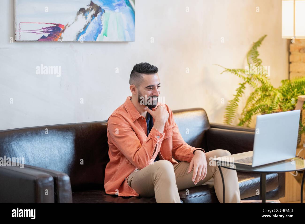 Happy young man sitting on sofa in front of laptop and communicating with teacher during online lesson Stock Photo