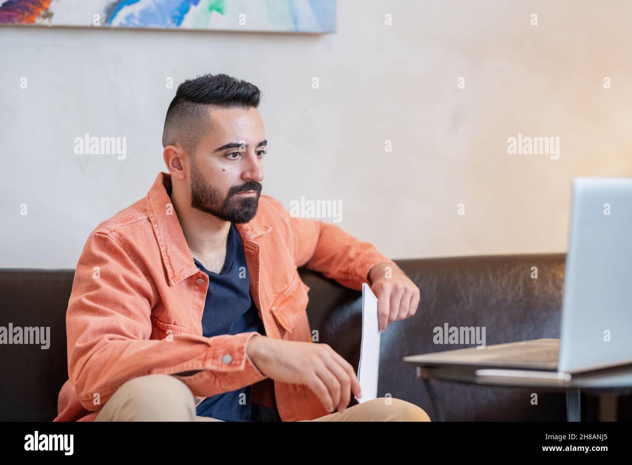 Young serious man looking at laptop screen while sitting on sofa in home environment during online lesson Stock Photo