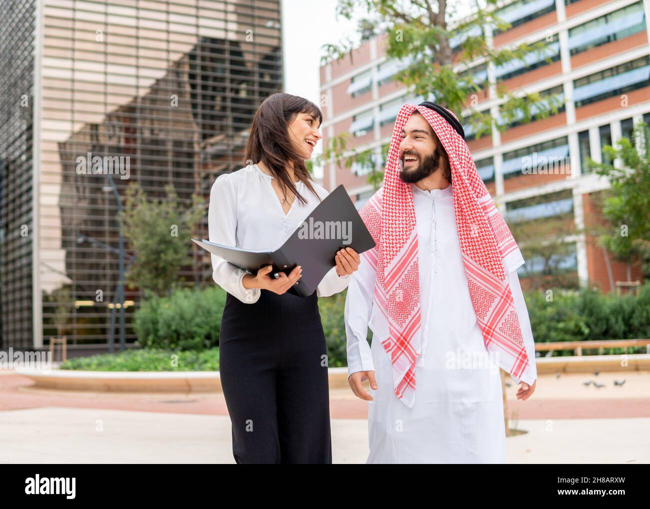 Diverse business people arab man and caucasian woman discussing documents while walking city street, young smiling business woman and arab man client negotiating contract at outdoor meeting Stock Photo