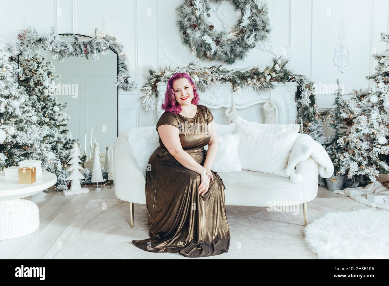 Beautiful young Caucasian plus size model woman in long golden olive color dress sitting on couch celebrating Christmas or New Year winter traditional Stock Photo