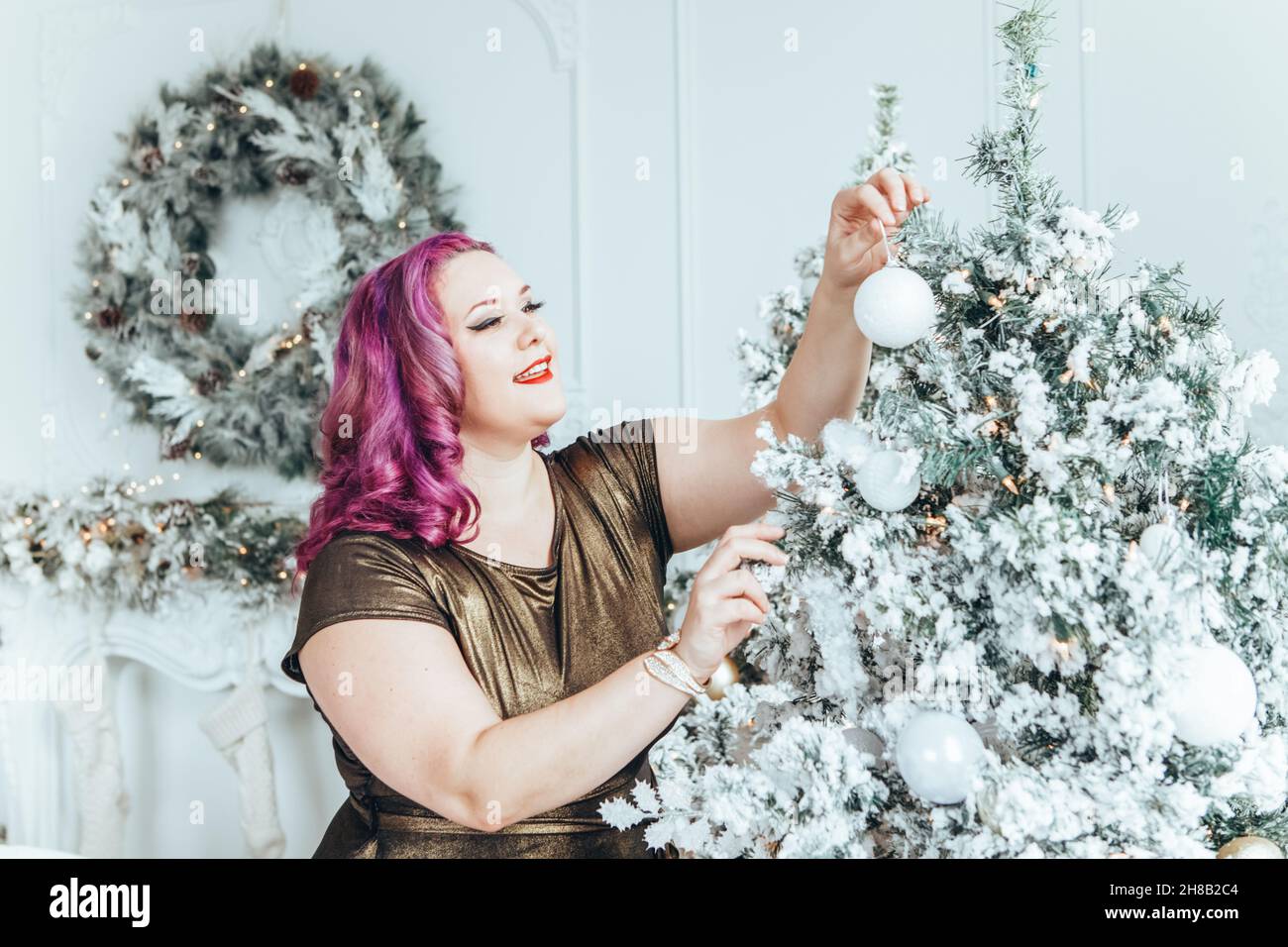 Beautiful young Caucasian plus size model woman in golden olive color dress decorating Christmas tree for holiday celebration at home.  Winter traditi Stock Photo