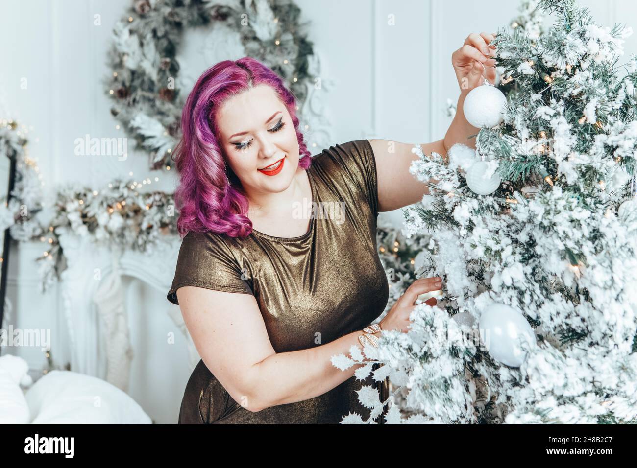 Beautiful young Caucasian plus size model woman in golden olive color dress decorating Christmas tree for holiday celebration at home.  Winter traditi Stock Photo