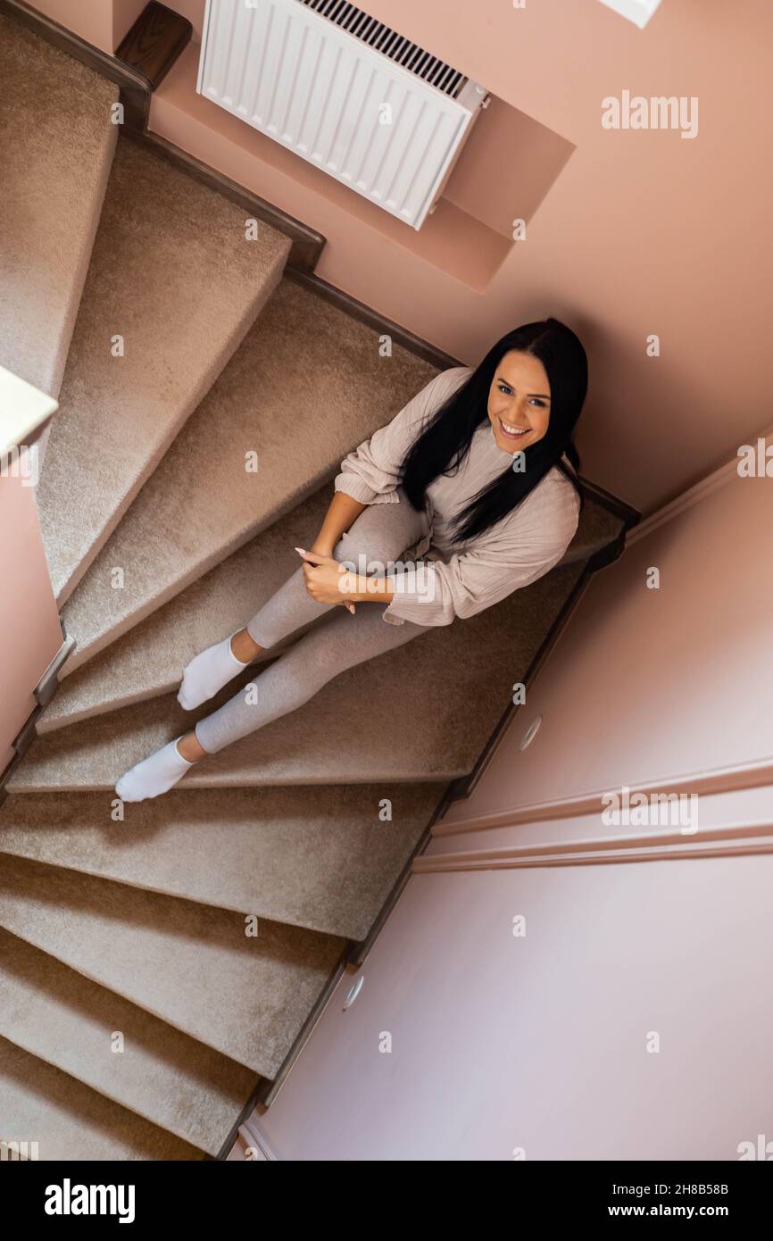 Top view happy young brunette casual woman wearing comfortable domestic outfit sitting on stairs Stock Photo