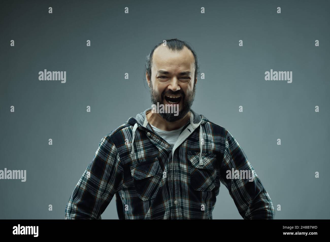 Wicked evil villainous man indulging in manic laughter as he faces the camera with hands on hips over a grey background with vignette Stock Photo
