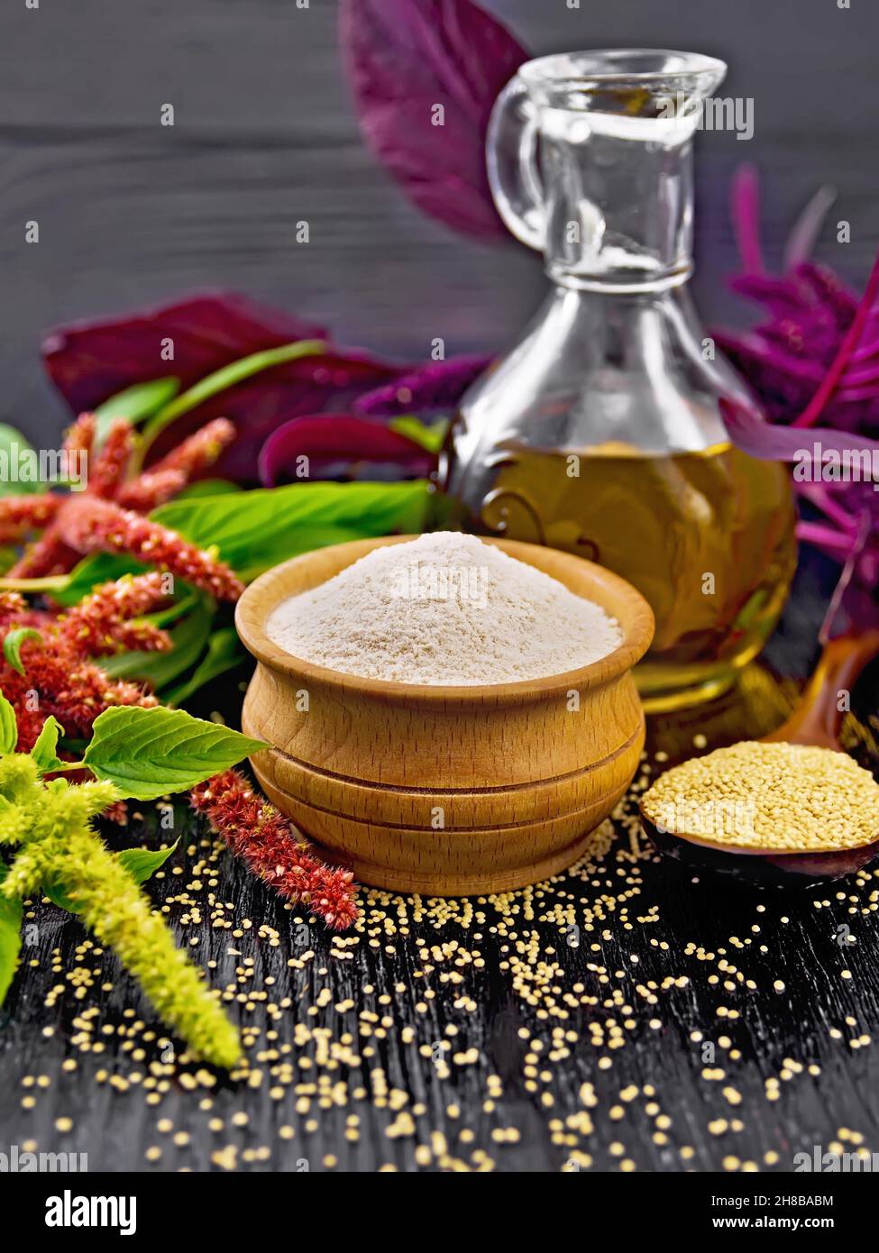 Amaranth flour in a bowl, seeds in a spoon and oil in decanter, leaves and brown, green, purple flowers of plant on wooden board background Stock Photo