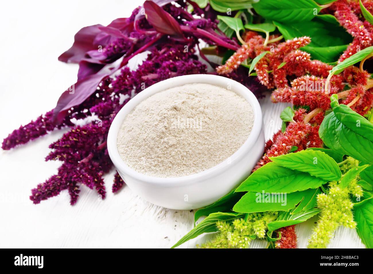 Amaranth flour in a bowl, brown, green and purple flowers and leaves of a plant on white wooden board background Stock Photo