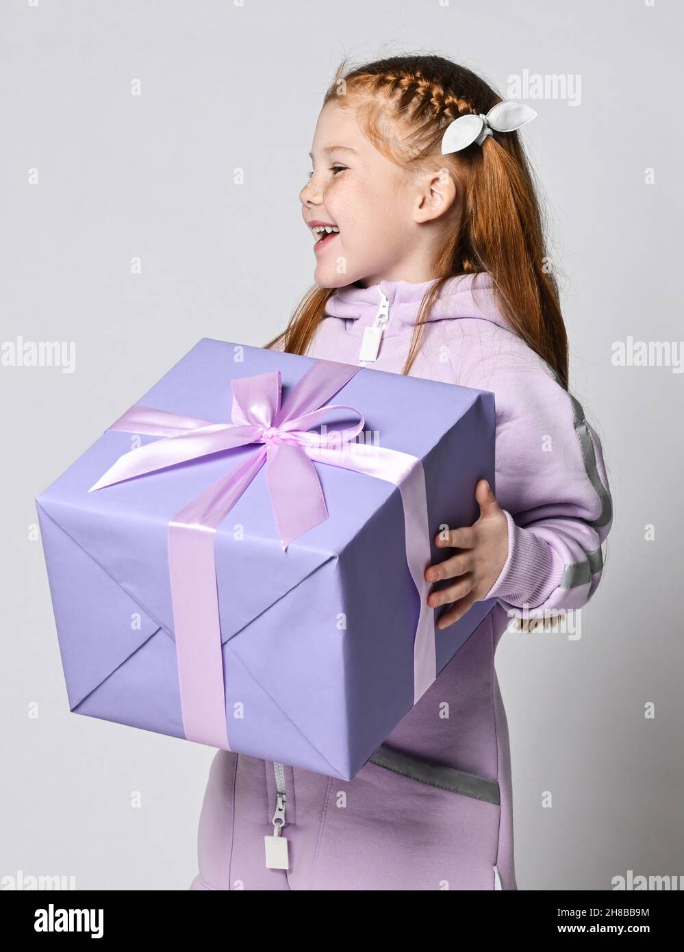 Portrait of happy laughing kid girl in pink jumpsuit with big blue gift box with ribbon and bow in hands looking aside Stock Photo
