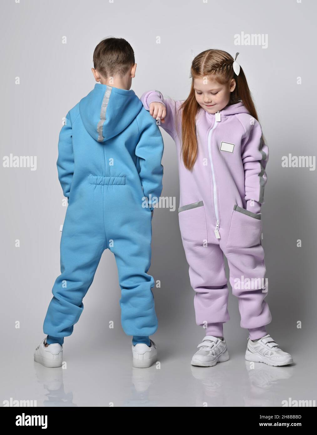 Two kids in jumpsuits are standing next to each other, boy with his back to us and girl with her face and looks down Stock Photo