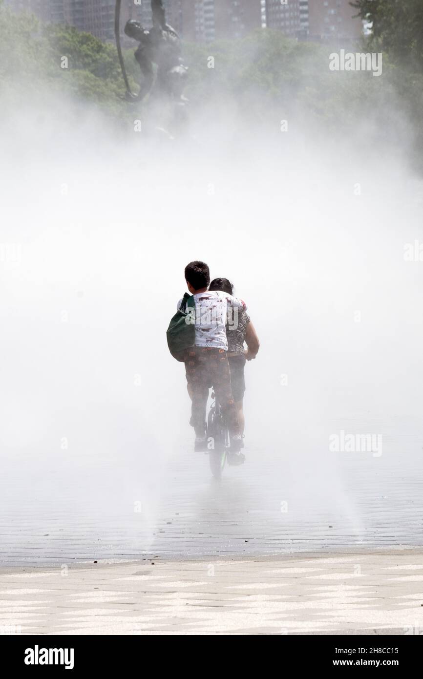 A mother & son on a bicycle pedal into a mist fountain near the Unisphere in Flushing Meadows Corona Park in Queens, New York. Stock Photo
