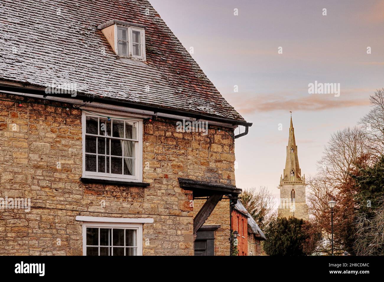Sharnbrook High Street, Bedfordshire, England, UK - Winter scene at sunrise of old cottage with frost on roof and distant view of village church spire Stock Photo