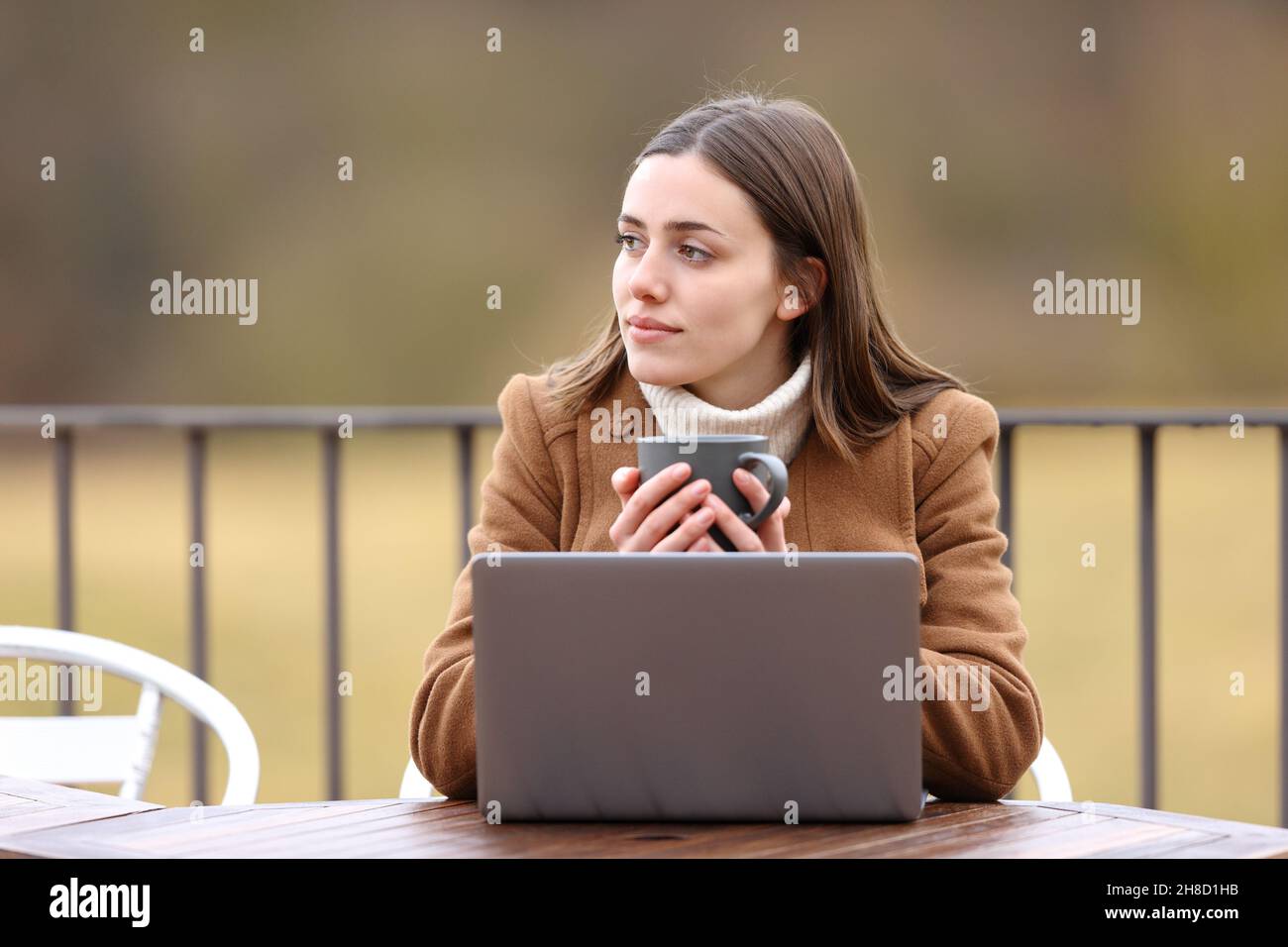 Relaxed woman with a laptop looking away holding coffee mug in a terrace in winter Stock Photo
