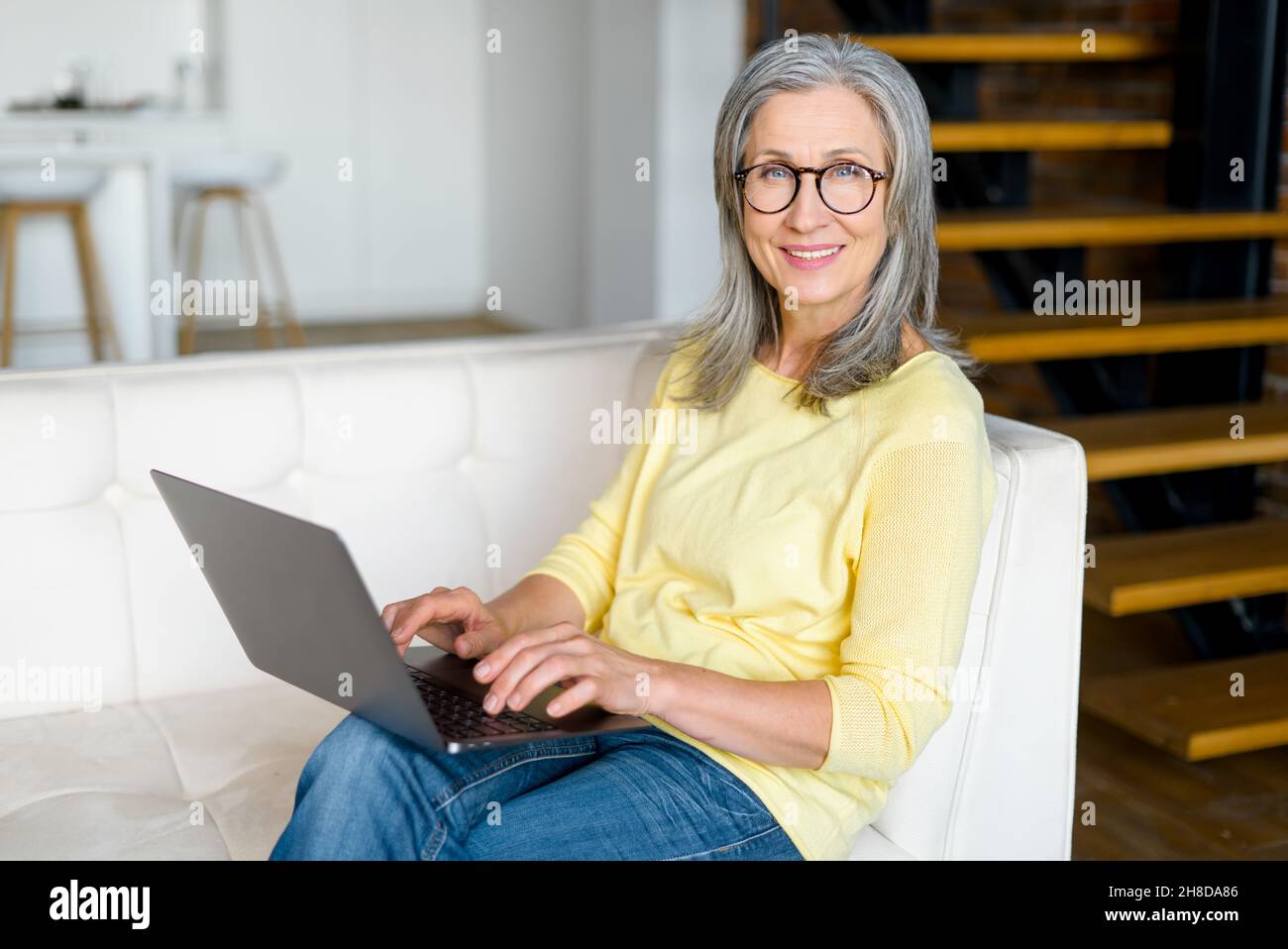 Positive charming mature gray-haired woman in glasses sitting on the sofa, typing on laptop, doing online shopping, working on a project, watching webinar. Freelancer enjoying remote work from home Stock Photo