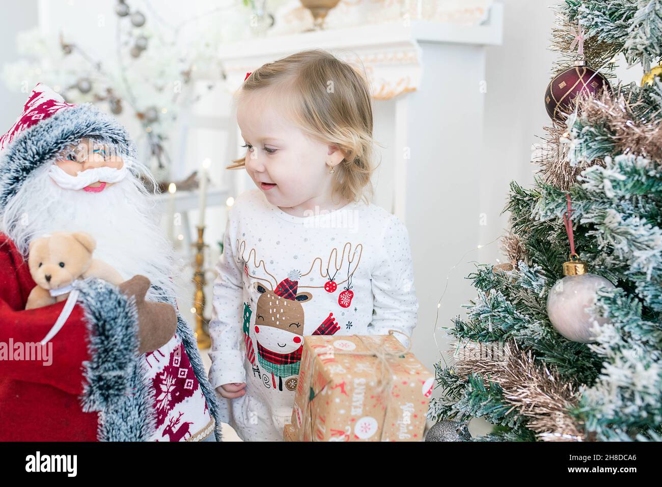 Adorable blonde little girl wearing festive pajamas while smiling and playing with the multitude of toys surrounding the decorated Christmas tree Stock Photo