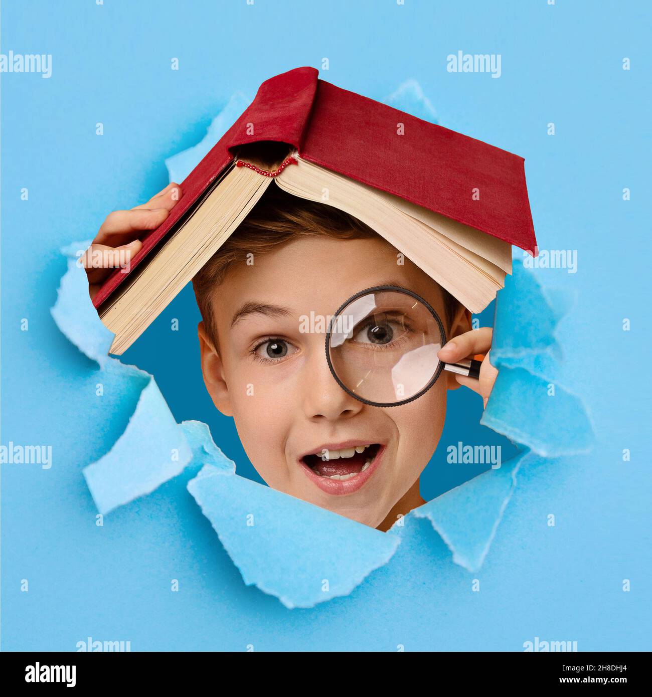 Cute clever boy with magnifying glass hiding under book, looking through hole in torn blue paper Stock Photo