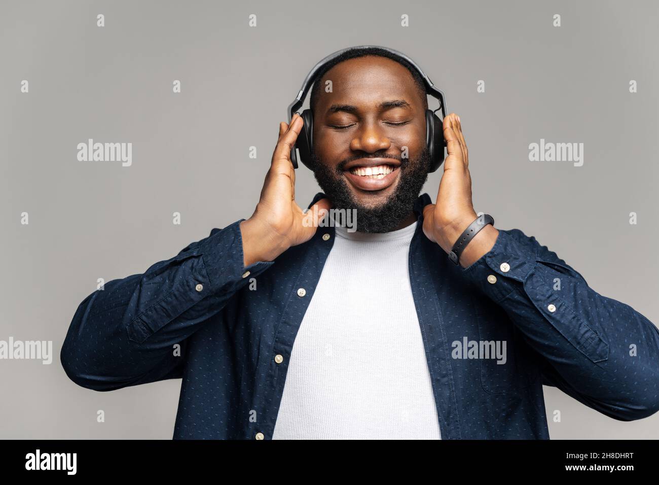 Studio portrait of relaxed cheerful African American man wearing and holding wireless headphones isolated on gray wall, enjoying listening to favorite music soundtrack, wellbeing, meditation concept Stock Photo