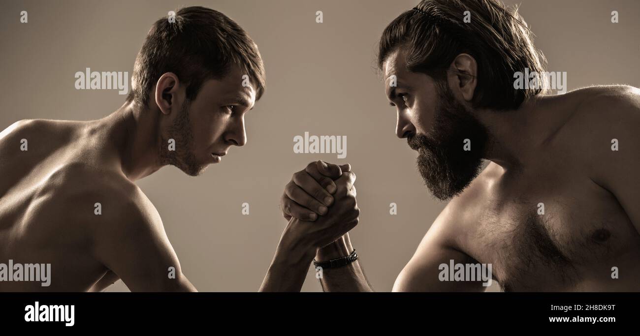 Heavily muscled bearded man arm wrestling a puny weak man. Arms wrestling thin hand, big strong arm in studio. Two man's hands clasped arm wrestling Stock Photo
