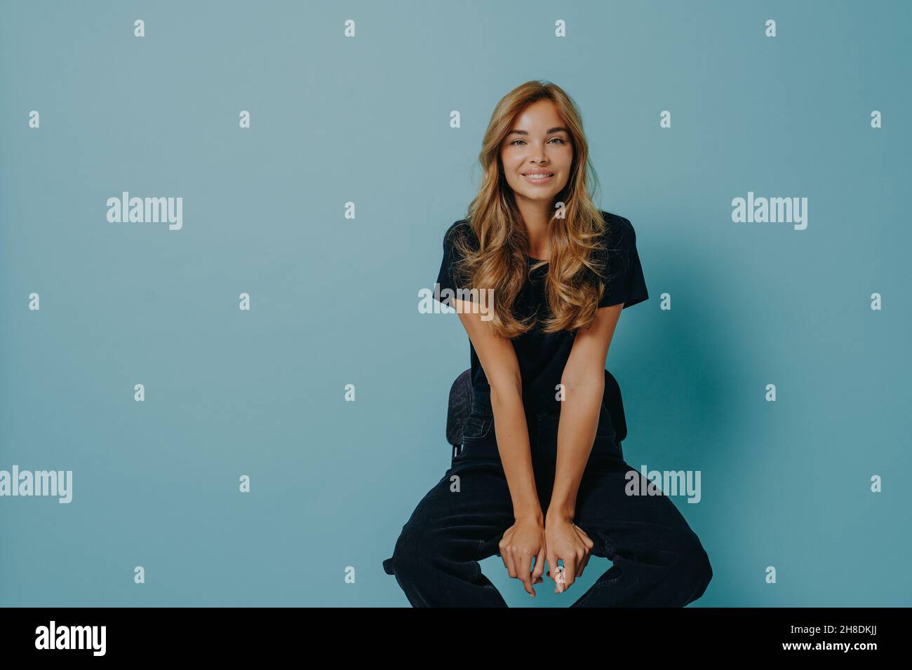 Beautiful young smiling lady hair in black casual t-shirt and jeans sitting on chair in studio Stock Photo