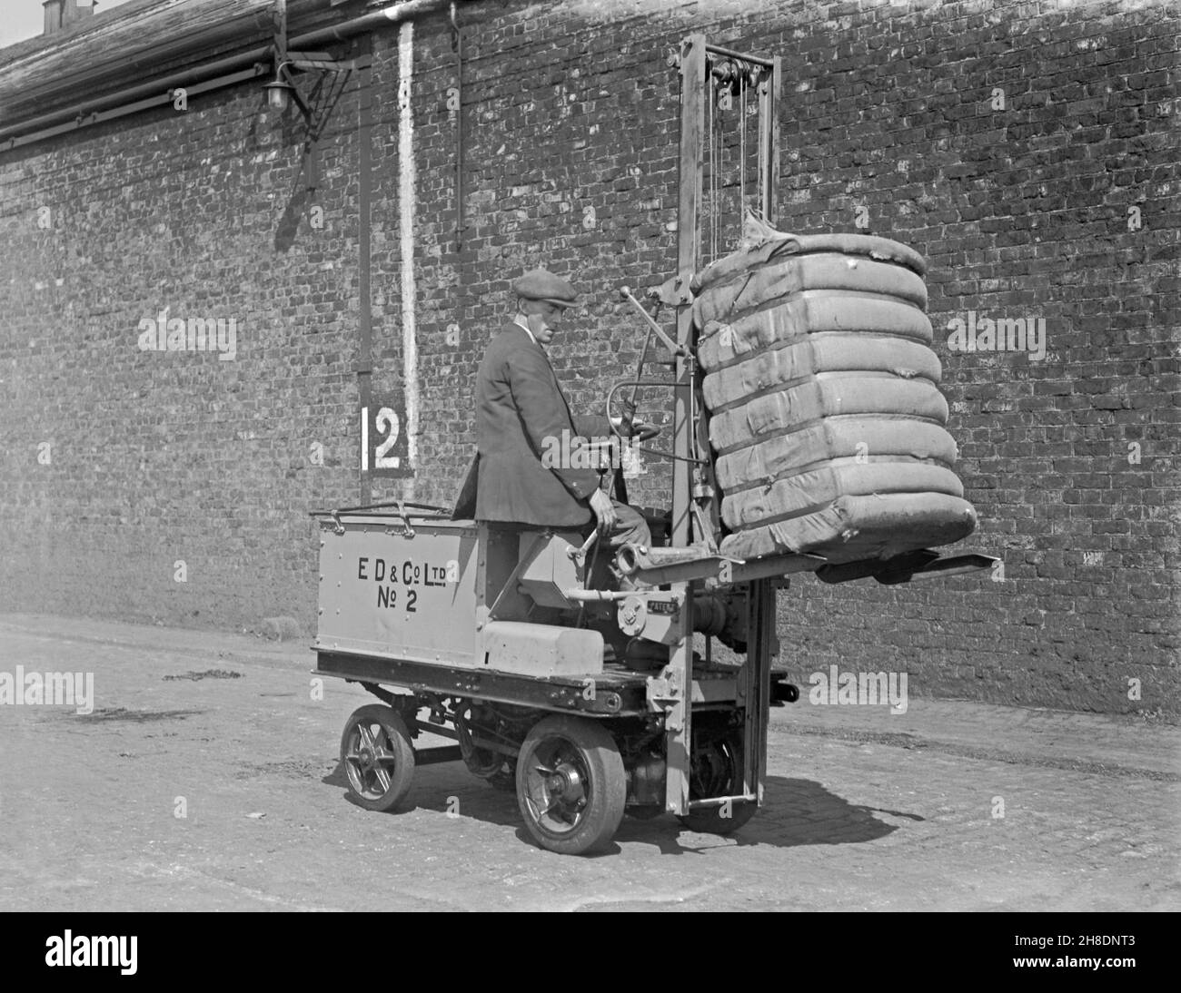 Early battery powered transport – here a forklift truck is being used to transport a heavy bale at E D and Co Ltd, a shipping and warehousing company at Liverpool Docks, Lancashire, England, UK in the early years of the twentieth century. In the early years of motor transport electrically-powered vehicles were a good option compared with petrol driven ones for short-distance commercial transport, such as moving goods around factories. This is taken from an old original glass negative – a vintage 20th century photograph. Stock Photo