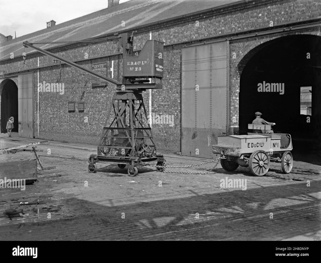 Early battery powered transport – here a Walker truck is towing a small mobile crane at E D and Co Ltd, a shipping and warehousing company at Liverpool Docks, Lancashire, England, UK in the early years of the twentieth century. Walker Electric Trucks were battery-powered vehicles built from 1907 to 1942 in the USA. In the early years of motor transport electrical power was a good option compared with petrol for short-distance commercial transport, such as moving goods around factories or local deliveries – a vintage 20th century photograph. Stock Photo