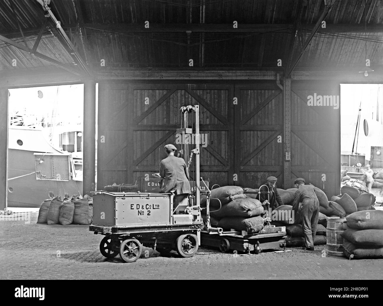 Early battery powered transport – here a forklift truck is being used to lift sacks on to a mobile weighing machine at E D and Co Ltd, a shipping and warehousing company at Liverpool Docks, Lancashire, England, UK in the early years of the twentieth century. In the early years of motor transport electrically-powered vehicles were a good option compared with petrol driven ones for short-distance commercial transport, such as moving goods around factories. This is taken from an old original glass negative – a vintage 20th century photograph. Stock Photo