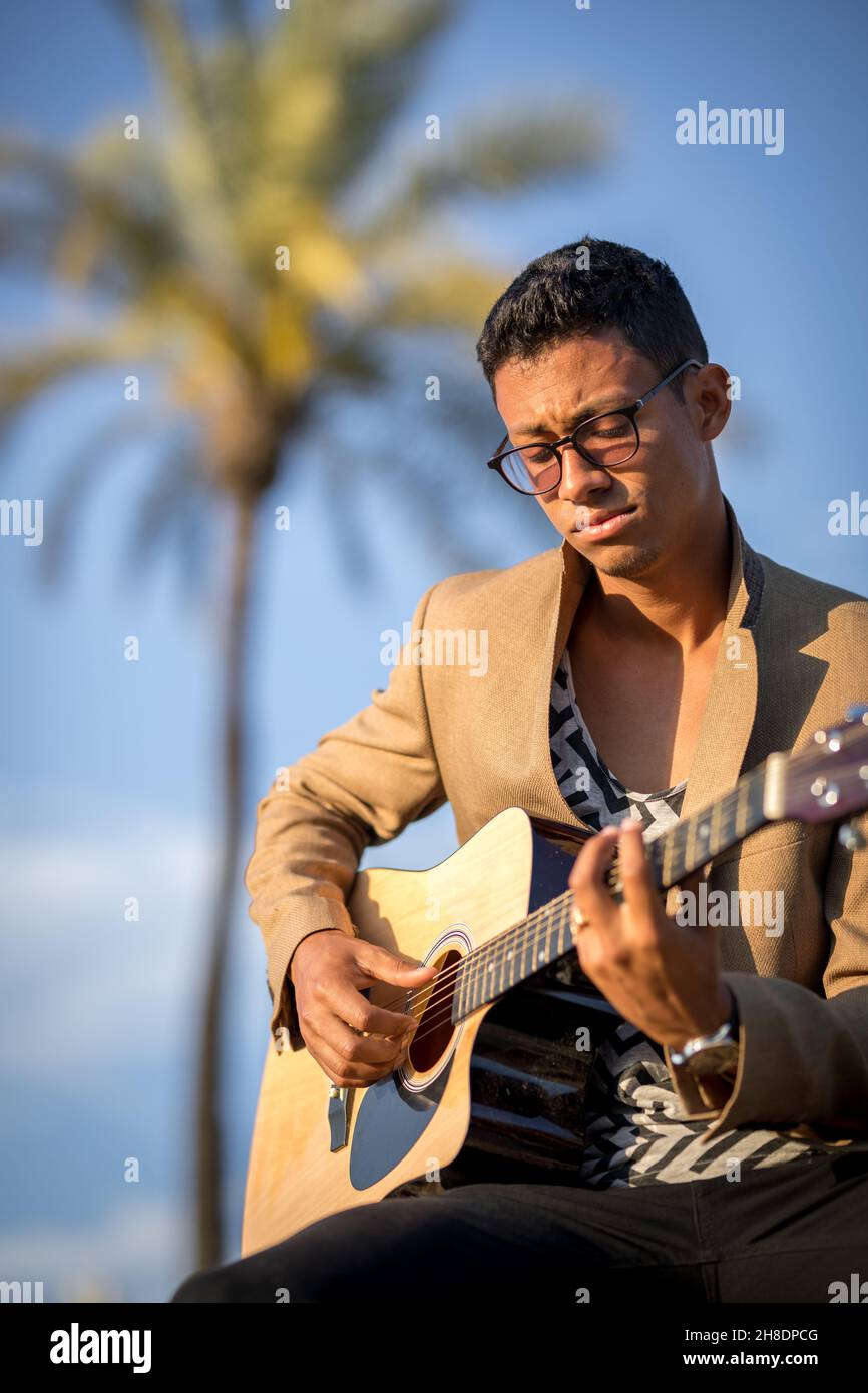 Portrait of Latin man sitting and playing guitar on street with palm trees in background Stock Photo